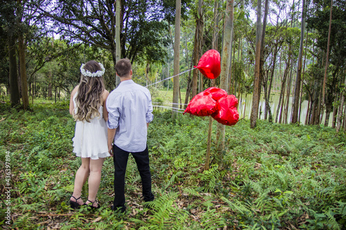 couple holding hands and back in their pre wedding in nature in a forest holding red heart balloons.