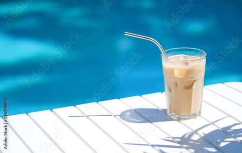 Ice coffee Cyprus Frappe Fredo against blue clear water of the swimming pool, on white table, with metal straw . Summer minimalistic background, holiday or vacation concept. Sun and shadows.Copy space photo