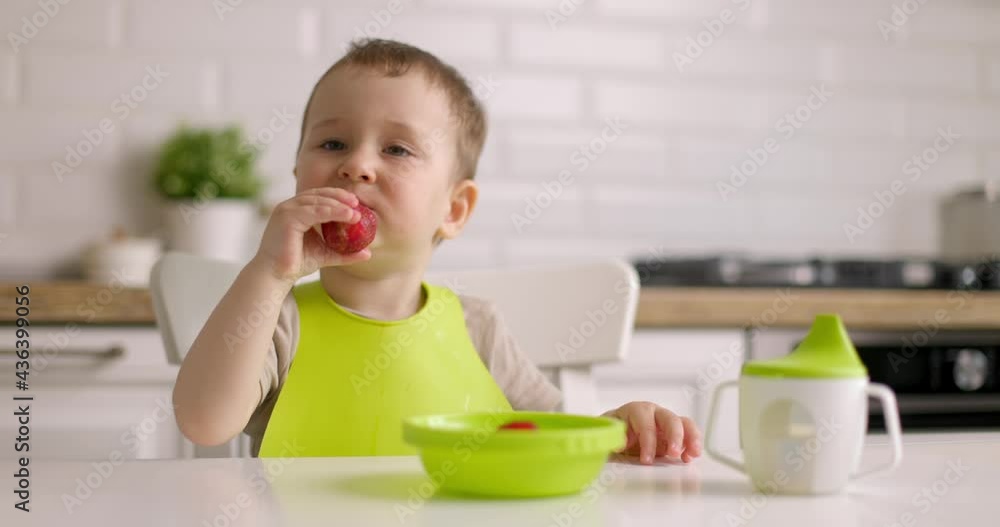 Cute baby boy sits at table in the kitchen and eats berries with his hand. The concept of happy childhood and healthy diet. Slow motion. Cute baby boy sits at table in the kitchen and eats berries with his hand. The concept of happy childhood and healthy diet. Slow motion.
