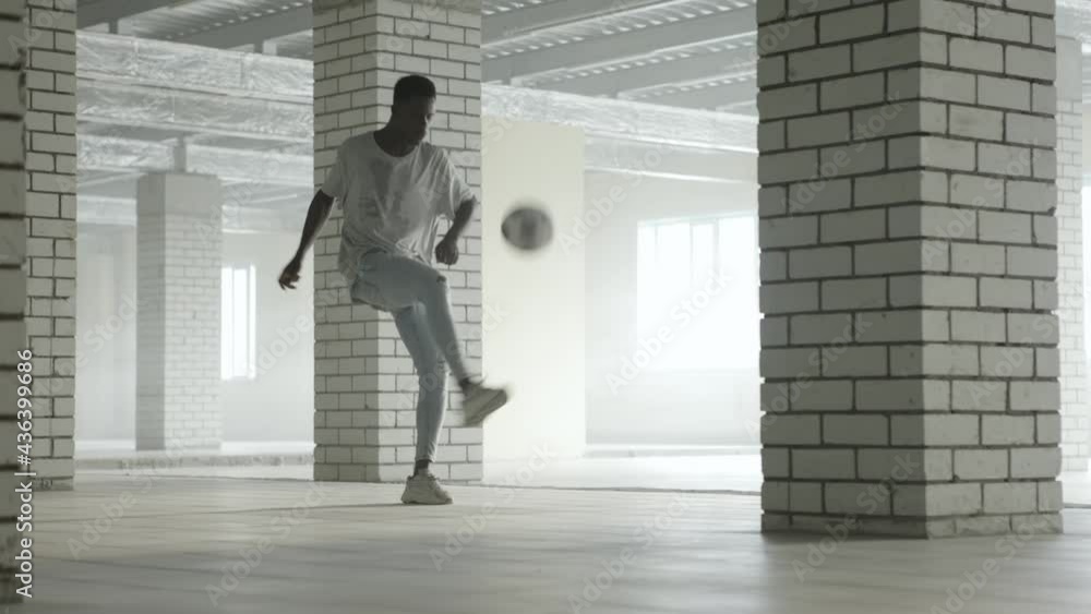 Young black guy trains with a soccer ball at indoors. Bright sunlight at background Young black guy trains with a soccer ball at indoors. Bright sunlight at background