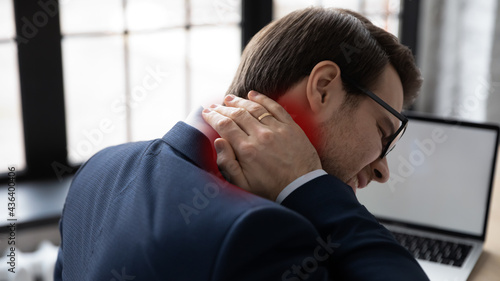 Frustrated young business man feeling strong neck ache after sitting at uncomfortable workplace, suffering from inflammation, osteochondrosis, muscles tension. Healthcare at work concept