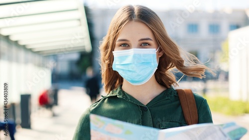 Close up portrait of cheerful Caucasian young woman traveller in medical mask standing outdoor in town holding map and looking at camera in good mood. Traveling during coronavirus pandemic