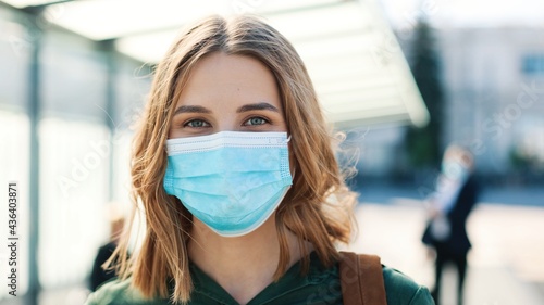 Close up portrait of cheerful Caucasian young woman in medical mask standing outdoor on sunny street on bus stop and looking at camera in good mood. Coronavirus pandemic, covid-19 concept