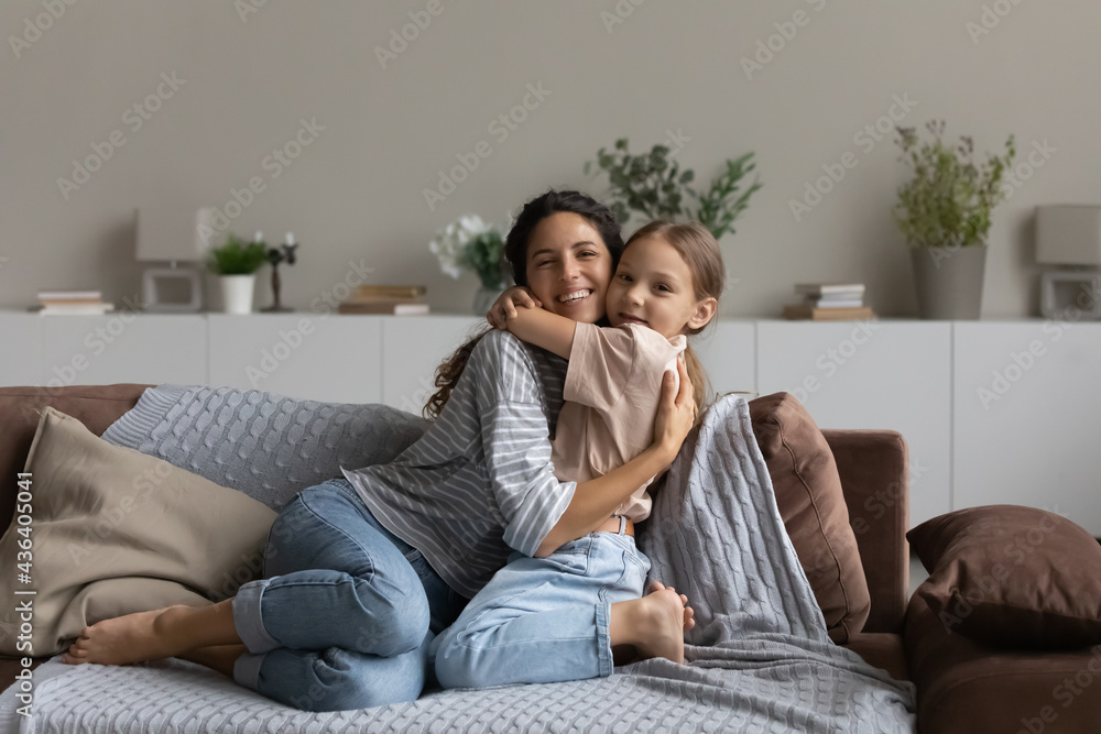 Portrait of smiling little biracial girl child hug young Hispanic mother relax together at home. Happy Latino mom and small daughter cuddle embrace together show love and care. Bonding concept.