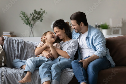 Happy young multiethnic family with small daughter relax on sofa hug cuddle enjoy weekend at home together. Smiling parents with little girl child sit rest on couch in living room. Rent concept.