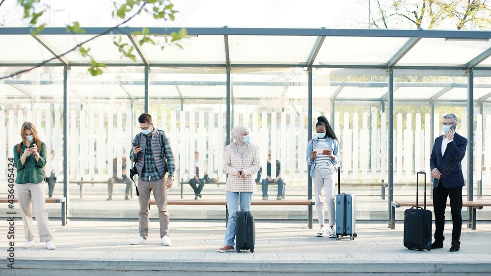 Different mixed-races people with masks on faces at bus stop or train station with suitcases keeping social distance. Man talking on cellphone. Woman texting on smartphone, travellers, Covid concept