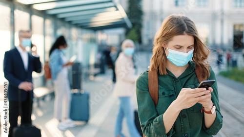 Close up of Caucasian beautiful young woman wearing medical mask and texting on smartphone browsing online standing outdoors on street at bus stop or station. Tourists waiting on background, covid-19