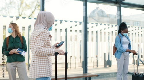 Portrait side view of Muslim woman traveller in medical mask with suitcase walking by bus stop and typing on cellphone gadget. Mixed-race tourists people on background. Covid-19 pandemic travel