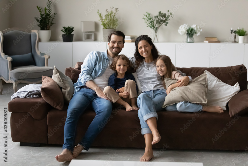 Portrait of smiling young multiracial family with two small kids sit relax on couch in living room. Happy multiethnic parents renters with little children rest at home together. Rental concept. Portrait of smiling young multiracial family with two small kids sit relax on couch in living room. Happy multiethnic parents renters with little children rest at home together. Rental concept.