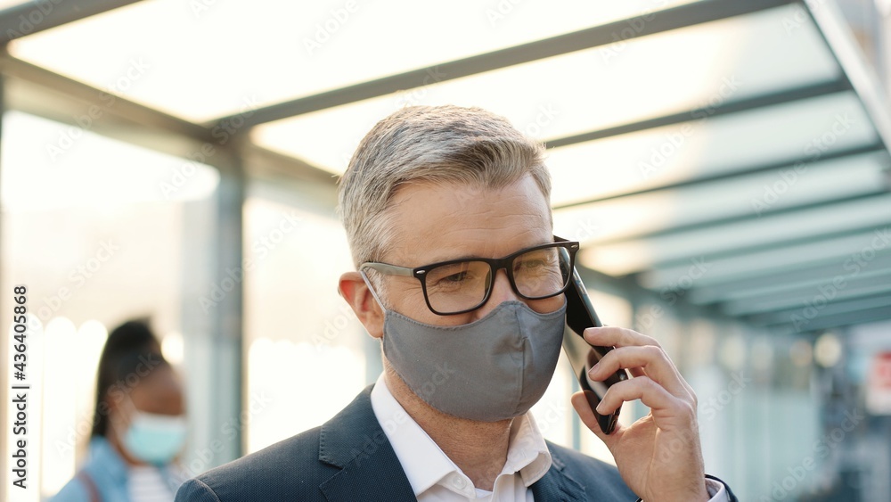 Close up of joyful positive adult senior main glasses wearing mask speaking on smartphone while standing in town on street. Happy handsome male calling on mobile phone in good mood outdoors Close up of joyful positive adult senior main glasses wearing mask speaking on smartphone while standing in town on street. Happy handsome male calling on mobile phone in good mood outdoors