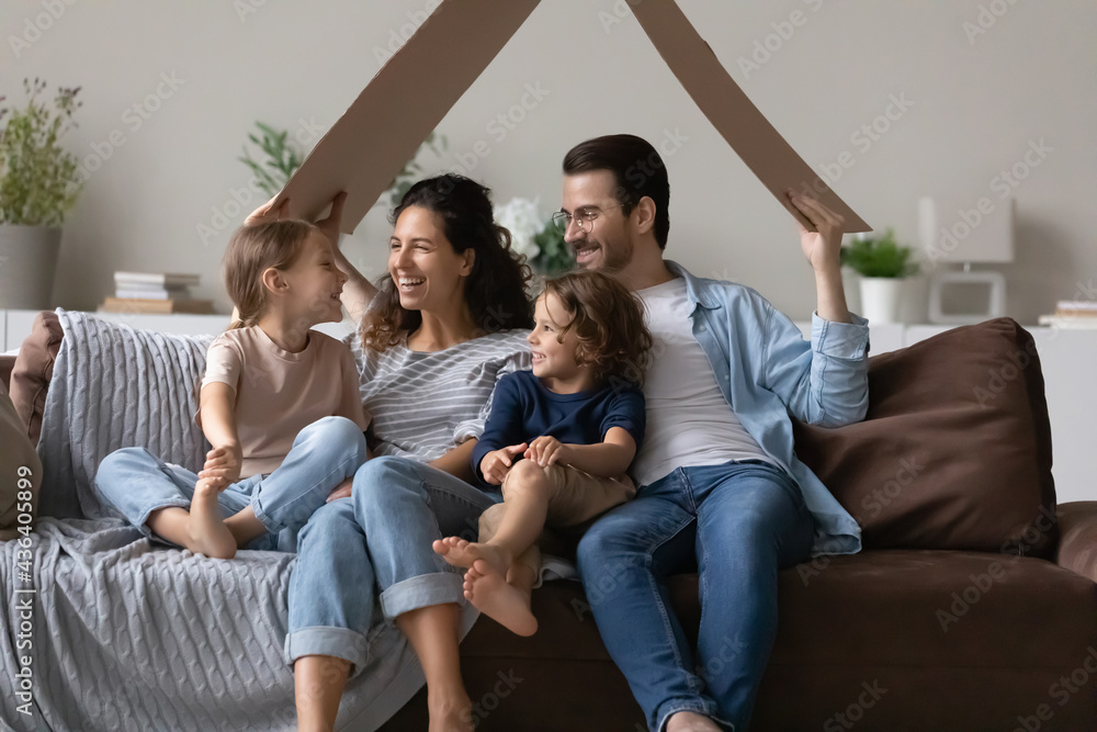 Overjoyed young family with small children relax in new own house excited moving relocating. Happy parents renters hold paper roof relax with kids at home, take bank loan mortgage. Rental concept. Overjoyed young family with small children relax in new own house excited moving relocating. Happy parents renters hold paper roof relax with kids at home, take bank loan mortgage. Rental concept.