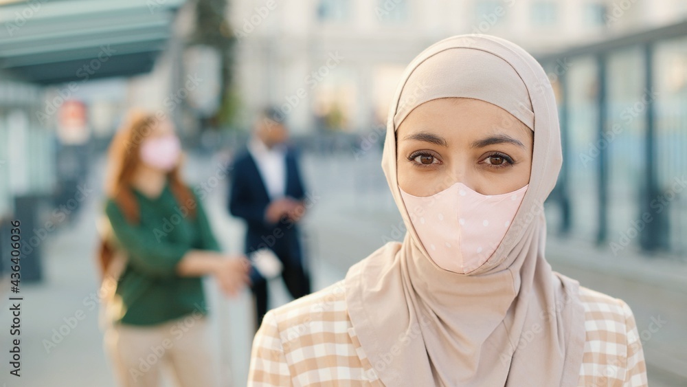 Close up of Muslim young beautiful female wearing mask on face looking at camera while standing in town at bus stop waiting outdoors. People travellers on background, coronavirus pandemic, new reality Close up of Muslim young beautiful female wearing mask on face looking at camera while standing in town at bus stop waiting outdoors. People travellers on background, coronavirus pandemic, new reality