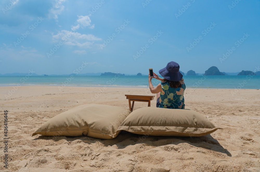 young woman sitting on swing near beach and using smartphone to take photo during summer vacation