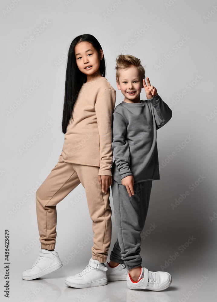 Fototapeta premium Caucasian kid boy and Asian kid girl friends in casual clothing pants, hoodies and sneakers stand together back to back over gray background