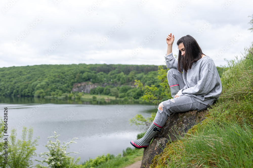 A girl in rubber boots sits on a stone near a lake in the forest.