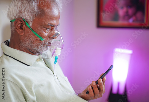 Old man with Oxygen concentrator mask reading news using mobile phone during home isolation - Concept of low oxygen level or therapy treatment from home for covid-19 coronavirus infected people photo