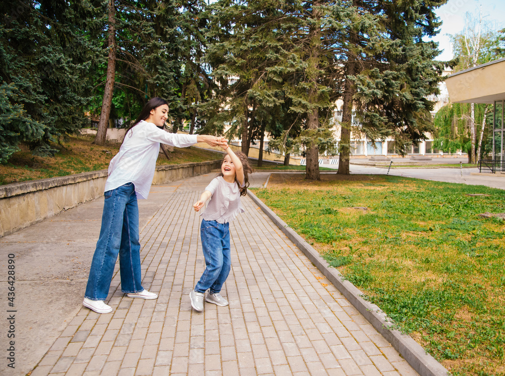 A young mother and her little daughter are dancing in the park. A girl and a woman are circling on the sidewalk.