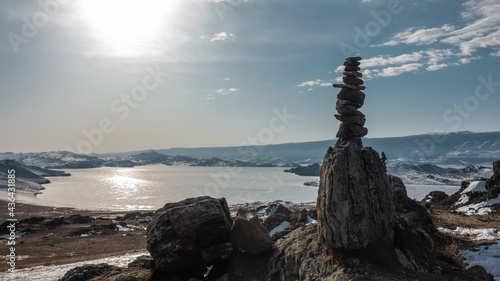 The frozen lake is surrounded by snow-capped mountains. Sun glare on ice. On the shore, against the background of the blue sky, a pyramid of stones is built. Baikal