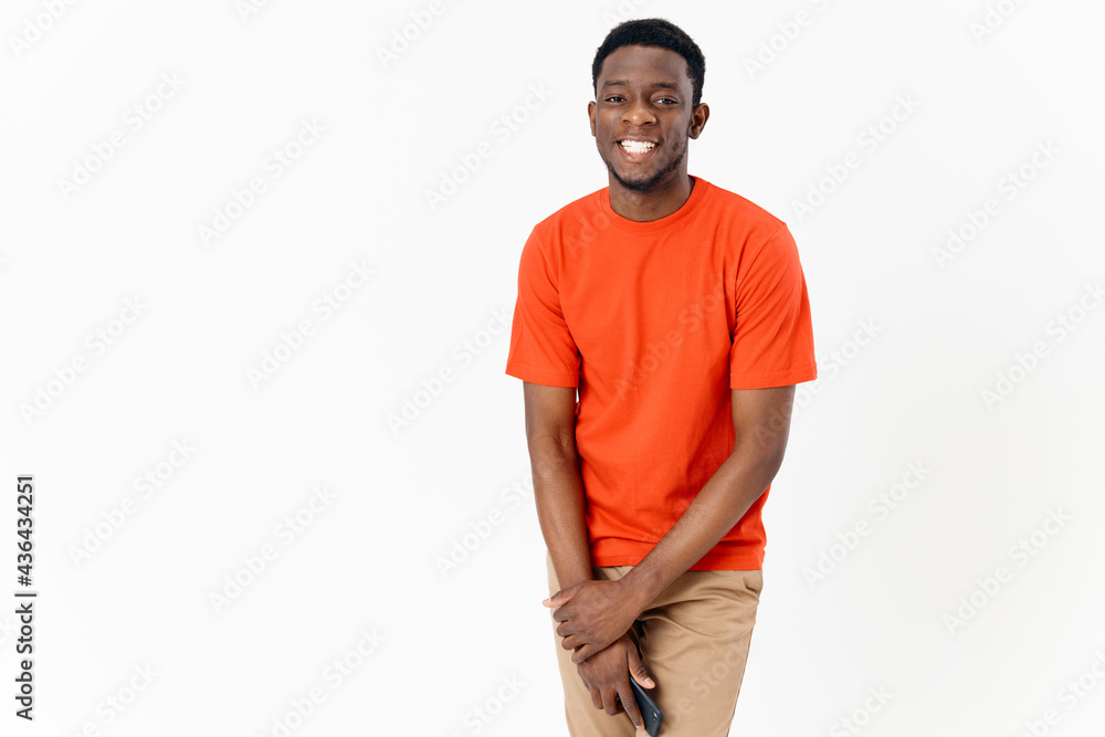 Fototapeta premium African guy in an orange T-shirt with a mobile phone and in trousers on a light background