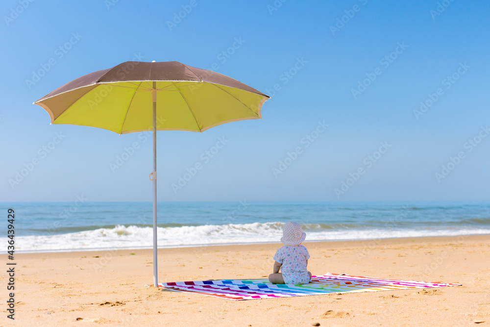 Baby sitting on his back on a colourful striped towel under an umbrella looking out at the beach.