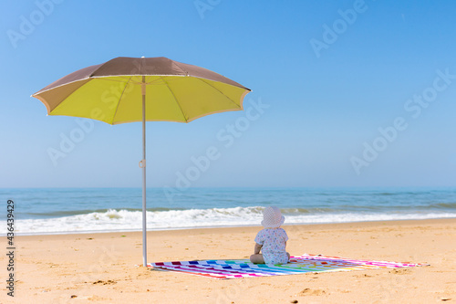 Fototapeta Naklejka Na Ścianę i Meble -  Baby sitting on his back on a colourful striped towel under an umbrella looking out at the beach.