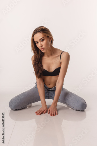portrait of sexy asian woman with long hair posing in black lingerie and blue jeans on white studio background. model tests of skinny girl in bra. attractive female sitting on floor on her knees