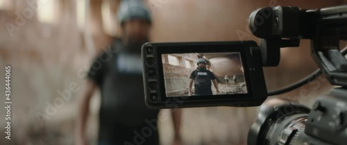 Behind the scenes of war journalist correspondent wearing bulletproof vest and helmet reporting live near destroyed building, military personnel in the background