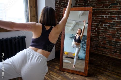 View from back of beautiful sporty woman practicing yoga, standing in Natarajasana exercise Lord Dance pose in front of the mirror in loft, looking at your reflection, lifestyle healthy concept. 