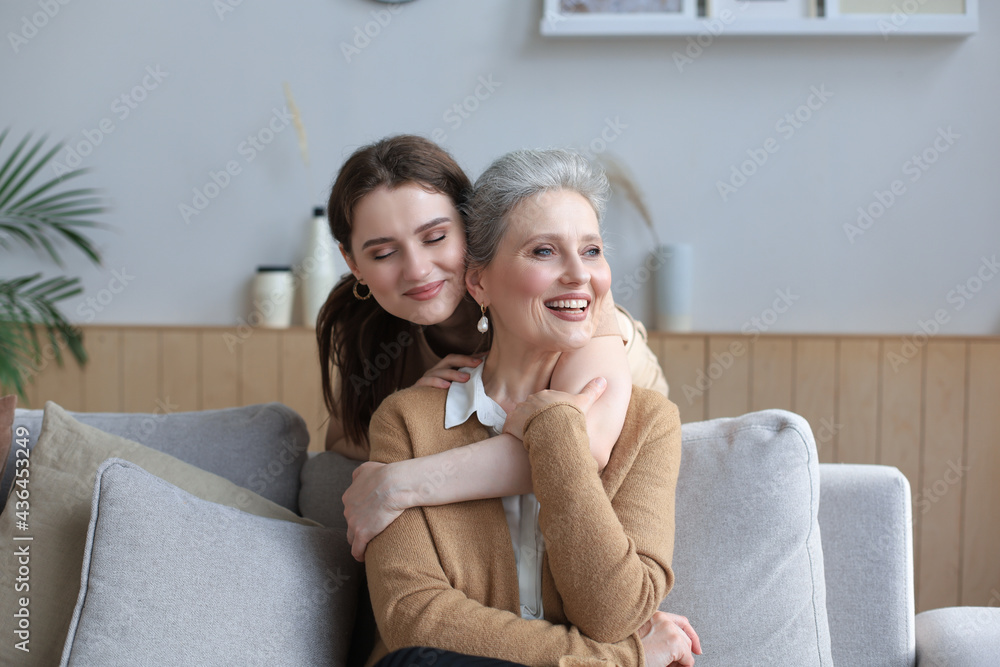 Obraz premium Happy daughter hugging older mother, standing behind sofa in living room, enjoying tender moment at home