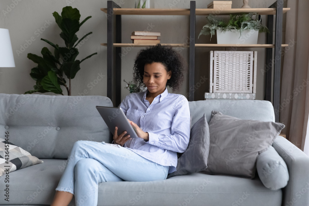Happy young African American woman sit relax on couch at home use modern tablet gadget. Smiling millennial biracial female rest on sofa in living room talk on webcam video call on pad device. Happy young African American woman sit relax on couch at home use modern tablet gadget. Smiling millennial biracial female rest on sofa in living room talk on webcam video call on pad device.