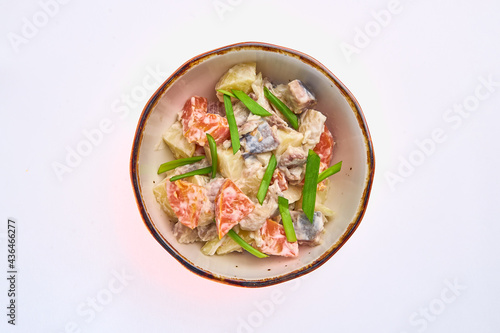 Salad of boiled potatoes, fresh tomatoes, salted herring fish with white sauce, decorated with green onion feathers on top in a white round plate on a white background.