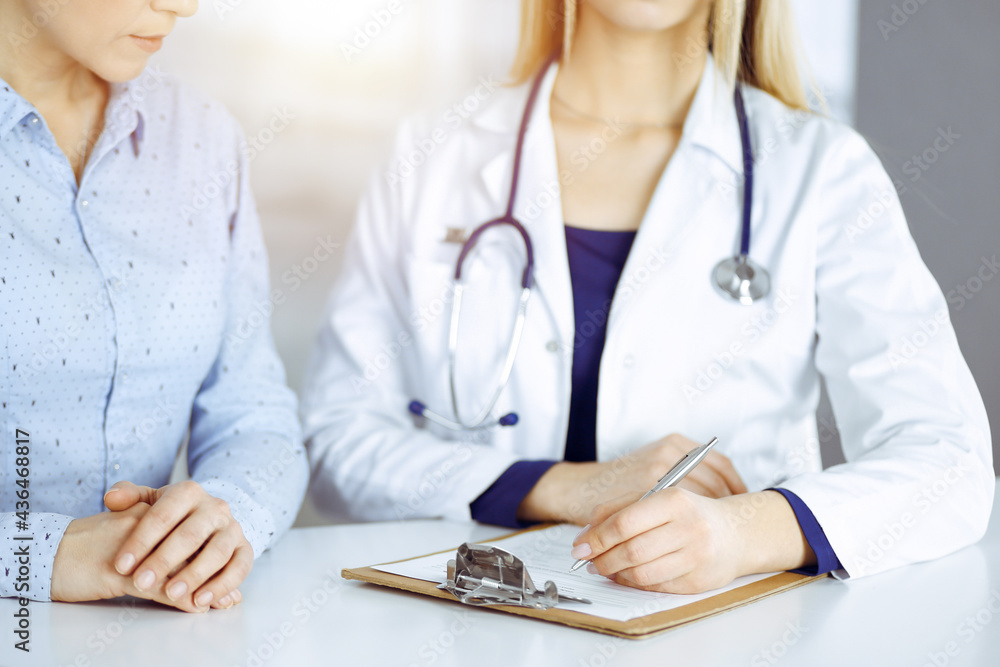 Young beautiful woman-doctor and her patient are discussing patient's current health examination, while sitting together at the desk in the sunny cabinet in a clinic. Female physician is writing some Young beautiful woman-doctor and her patient are discussing patient's current health examination, while sitting together at the desk in the sunny cabinet in a clinic. Female physician is writing some