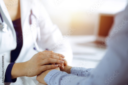 Unknown woman-doctor is holding her patient s hands  discussing current health examination  while they are sitting at the desk in the sunny cabinet in a clinic. Physician with a stethscope at work