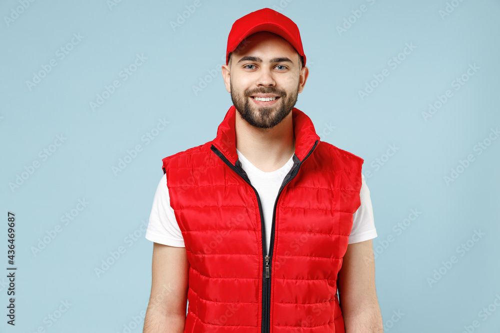 Professional happy delivery guy employee man in red cap white T-shirt vest uniform workwear work as dealer courier look camera isolated on pastel blue color background studio portrait Service concept