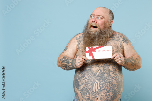 Young fun shocked fat pudge obese chubby overweight blue-eyed bearded man 30s has big belly with naked tattooed torso hold gift voucher flyer mock up look aside isolated on pastel blue background photo