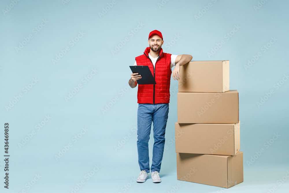 Full size body length delivery guy employee man in red cap white T-shirt vest uniform work as dealer courier hold blank cardboard box isolated on pastel blue color background studio. Service concept. Full size body length delivery guy employee man in red cap white T-shirt vest uniform work as dealer courier hold blank cardboard box isolated on pastel blue color background studio. Service concept.