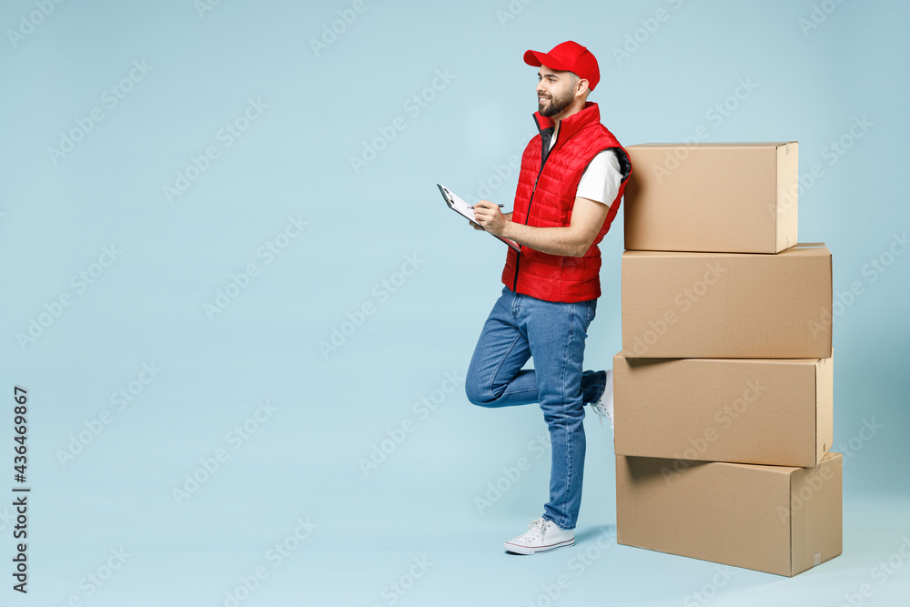 Full size body length delivery guy employee man in red cap white T-shirt vest uniform work as dealer courier hold blank cardboard box isolated on pastel blue color background studio. Service concept. Full size body length delivery guy employee man in red cap white T-shirt vest uniform work as dealer courier hold blank cardboard box isolated on pastel blue color background studio. Service concept.