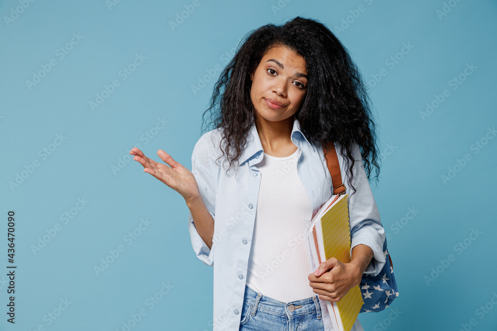 Young confused african american girl teen student wear denim clothes backpack hold books spread hands oops gesture isolated on blue background. Education in high school university college concept.