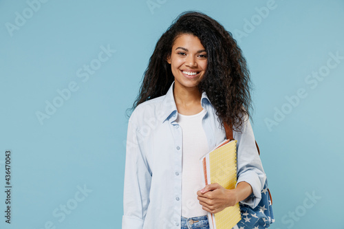 Young smiling happy african american girl teen student wear denim clothes backpack hold books isolated on blue color background studio portrait Fototapeta