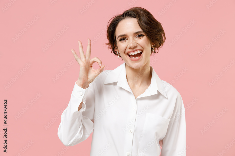 Young smiling happy successful employee business woman corporate lawyer 20s in classic formal white shirt work in office show ok okay gesture isolated on pastel pink color background studio portrait. Young smiling happy successful employee business woman corporate lawyer 20s in classic formal white shirt work in office show ok okay gesture isolated on pastel pink color background studio portrait.