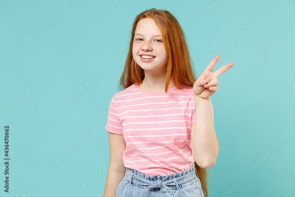 Little happy redhead kid girl 12-13 years old wearing pink striped t-shirt show victory v-sign gesture isolated on pastel blue background studio portrait. Children lifestyle childhood concept Little happy redhead kid girl 12-13 years old wearing pink striped t-shirt show victory v-sign gesture isolated on pastel blue background studio portrait. Children lifestyle childhood concept