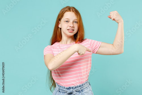 Little strong sporty redhead kid girl 12-13 years old in pink t-shirt point finger biceps muscles on hand demonstrating strength power isolated on pastel blue background. Lifestyle childhood concept