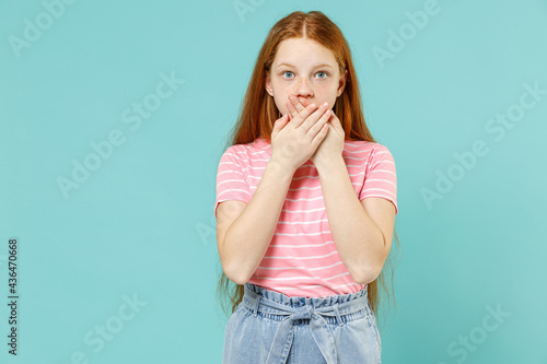 Little shocked surprised redhead kid girl 12-13 years old wear pink striped t-shirt cover mouth with hands arms isolated on pastel blue background studio portrait Children lifestyle childhood concept