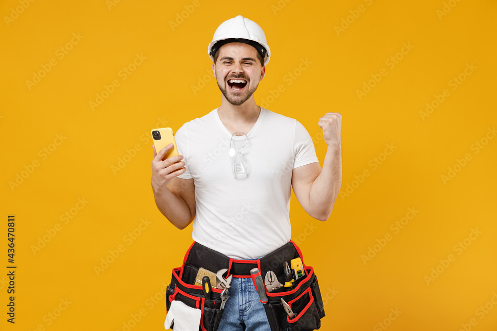 Young employee handyman man in protective helmet hardhat use mobile cell phone do winner gesture isolated on yellow background. Instruments accessories renovation apartment room. Repair home concept. Young employee handyman man in protective helmet hardhat use mobile cell phone do winner gesture isolated on yellow background. Instruments accessories renovation apartment room. Repair home concept.