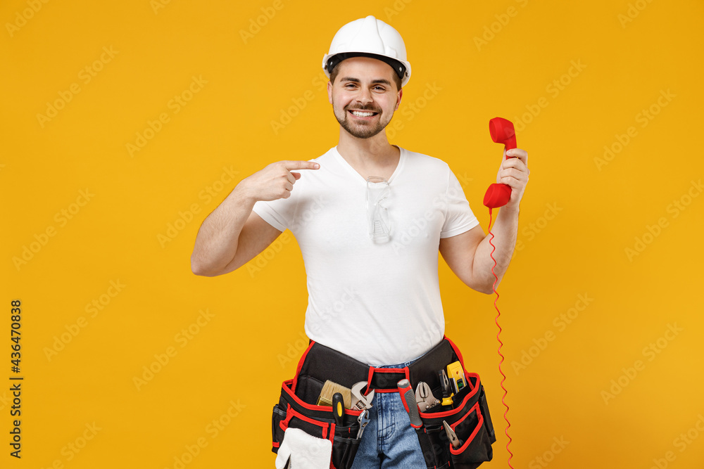 Young fun employee handyman man in protective helmet hardhat point finger on handset phone speak isolated on yellow background Instruments accessories renovation apartment room Repair home concept