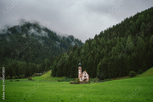 Church of St. Johann in Ranui on cloudy overcast day, Dolomite Alps, Vilnoss Valley, South Tyrol, Italy. Fog laxing over the pine tree forest photo