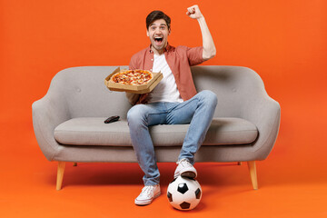 Young man football fan in shirt support favorite team with soccer ball sit on sofa at home watch tv live stream eat pizza do winner gesture isolated on orange background People sport leisure concept