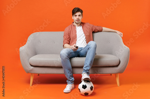 Young happy fun man football fan in shirt support favorite team with soccer ball sit on sofa at home watch tv live stream switch channel isolated on orange background. People sport leisure concept