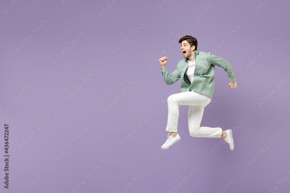 Full length side view excited amazed fun young man in casual mint shirt white t-shirt jump high running fast hurrying up look camera isolated on purple violet background. People lifestyle concept Full length side view excited amazed fun young man in casual mint shirt white t-shirt jump high running fast hurrying up look camera isolated on purple violet background. People lifestyle concept