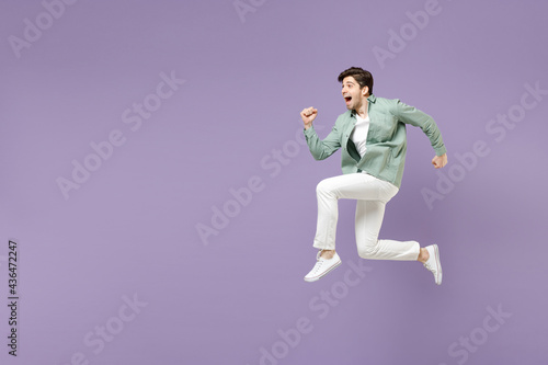 Full length side view excited amazed fun young man in casual mint shirt white t-shirt jump high running fast hurrying up look camera isolated on purple violet background. People lifestyle concept
